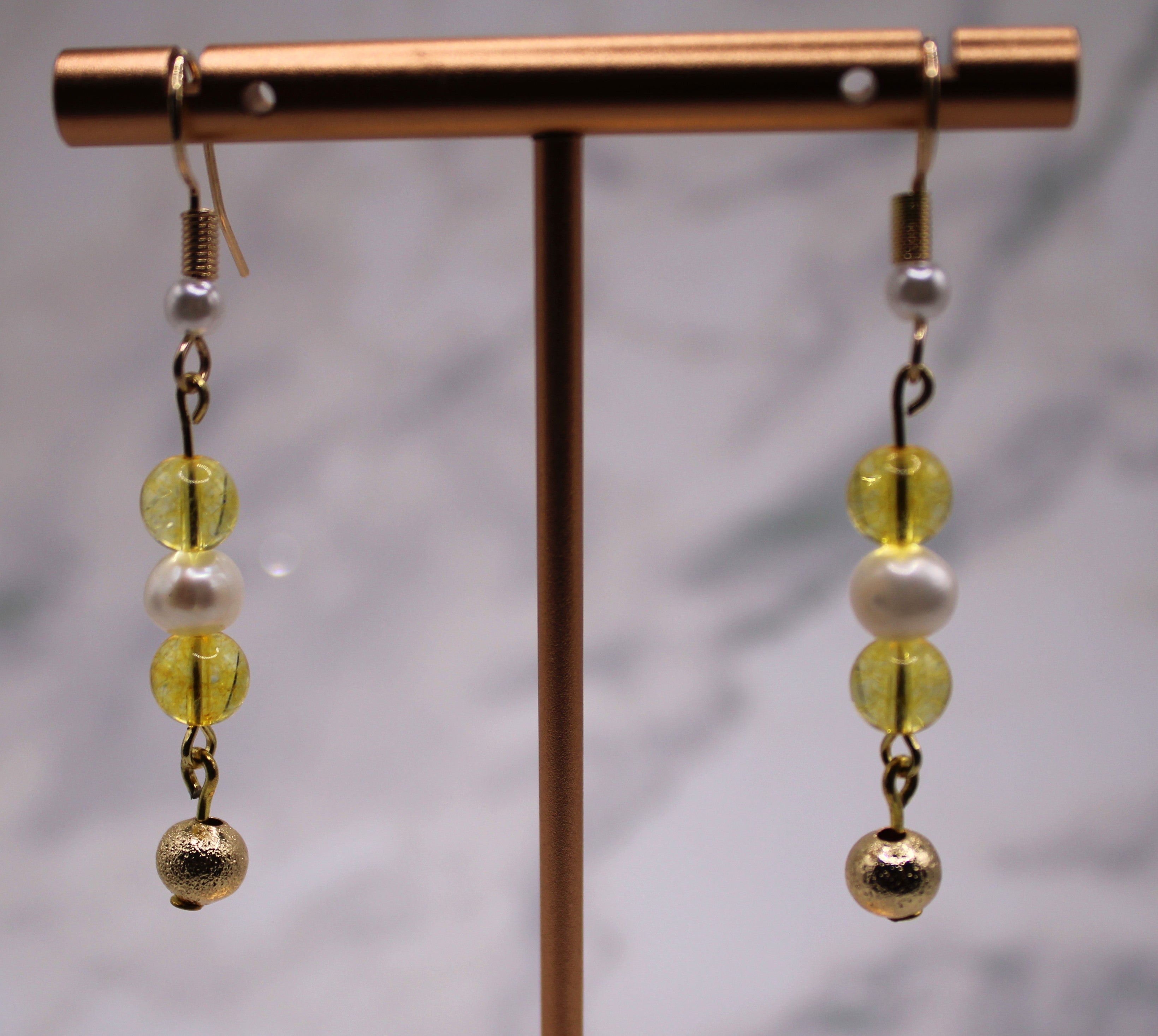A pair of dangling earrings displayed on a metallic stand. Each earring has a gold fishhook, a small white pearl bead, two translucent yellow beads, and a textured gold ball at the bottom.
