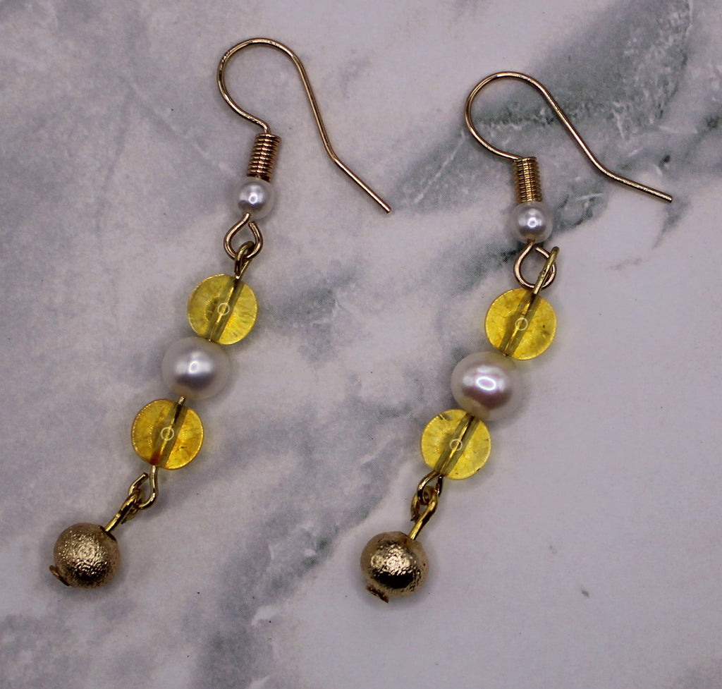 A pair of gold-tone dangle earrings with white pearl-like beads, translucent yellow disc beads, and small gold bells, displayed on a light gray marble surface.