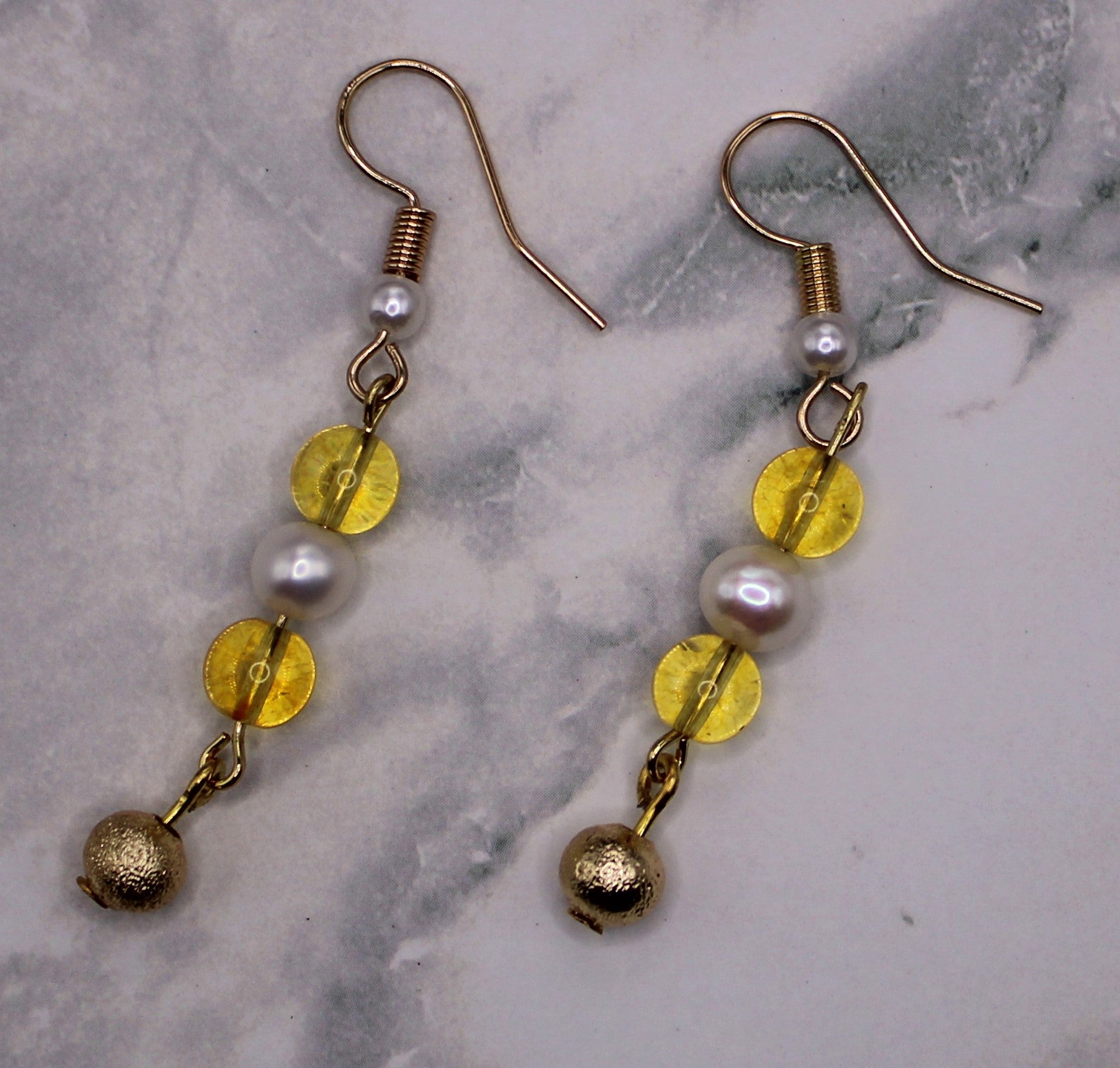 A pair of gold-tone dangle earrings with white pearl-like beads, translucent yellow disc beads, and small gold bells, displayed on a light gray marble surface.