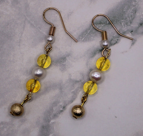 A pair of gold-tone dangle earrings with white pearl-like beads, translucent yellow disc beads, and small gold bells, displayed on a light gray marble surface.