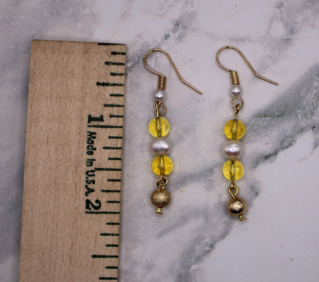 A pair of gold dangle earrings with yellow beads, white faux pearls, and a gold ball charm, displayed next to a wooden ruler on a white and gray marble surface.