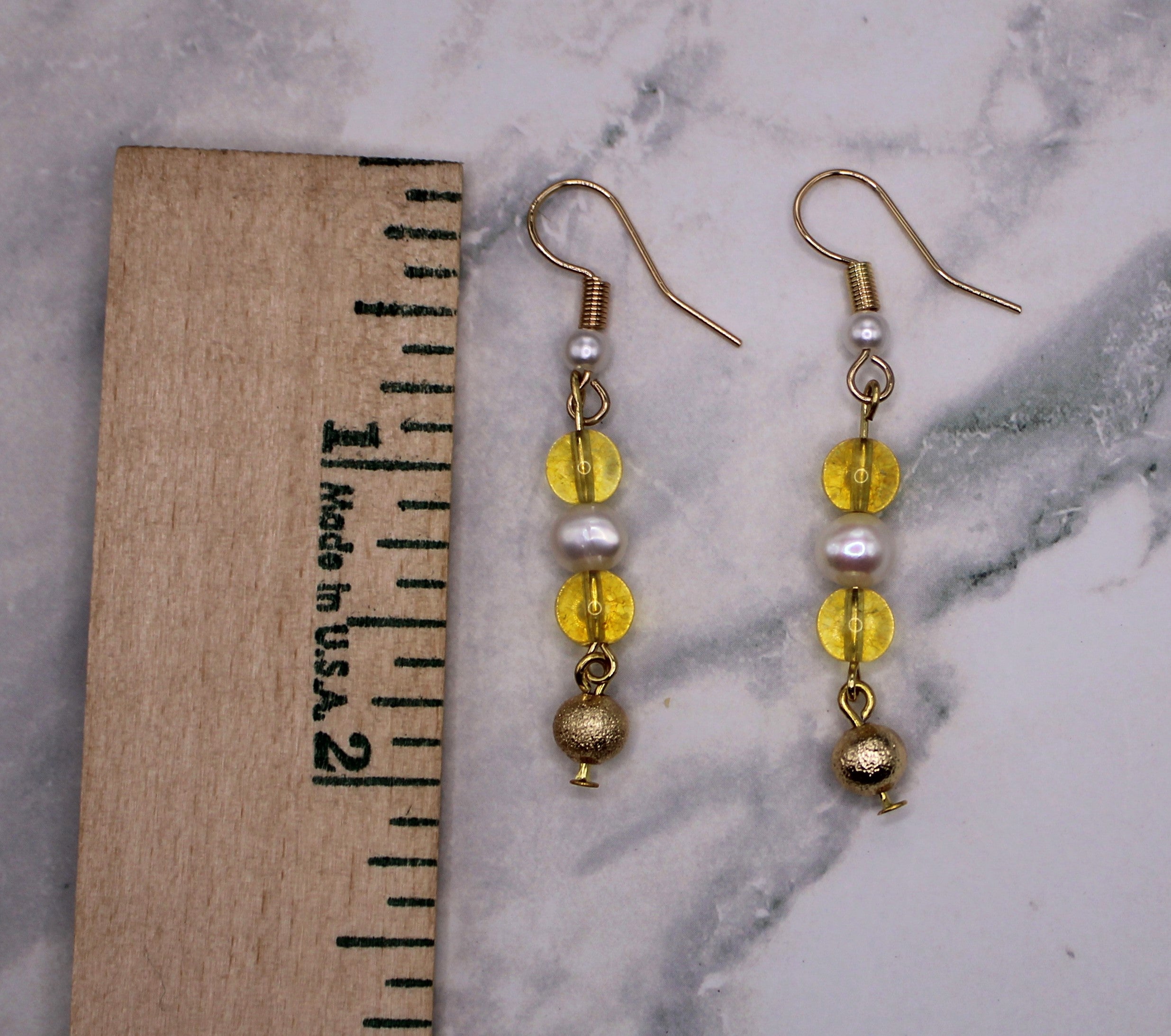A pair of gold dangle earrings with yellow beads, white faux pearls, and a gold ball charm, displayed next to a wooden ruler on a white and gray marble surface.