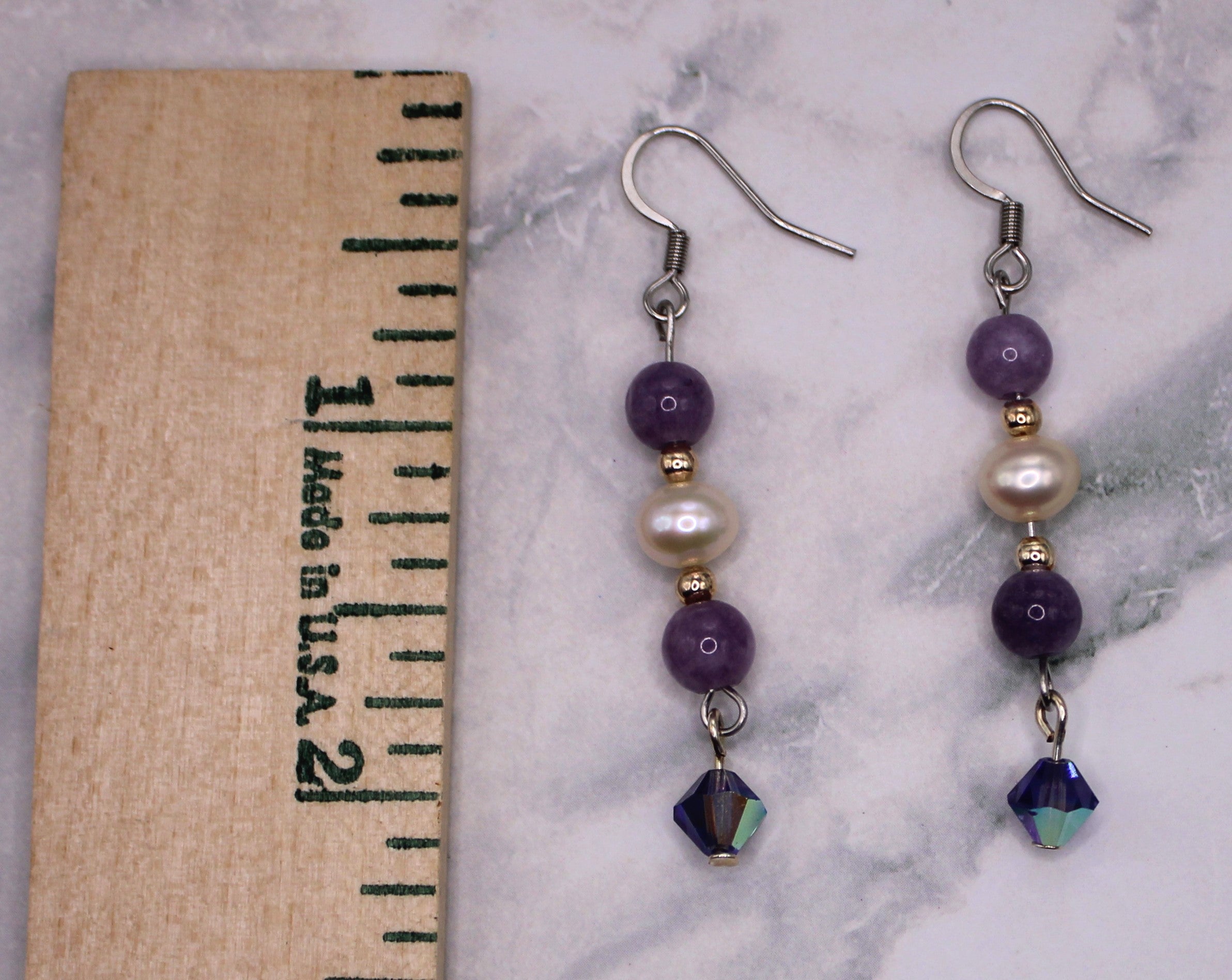 A pair of handmade dangle earrings displayed on a white marble surface next to a wooden ruler for scale. The earrings feature purple beads, a white pearl, a small gold spacer bead, and a faceted dark blue-green iridescent bead at the bottom.