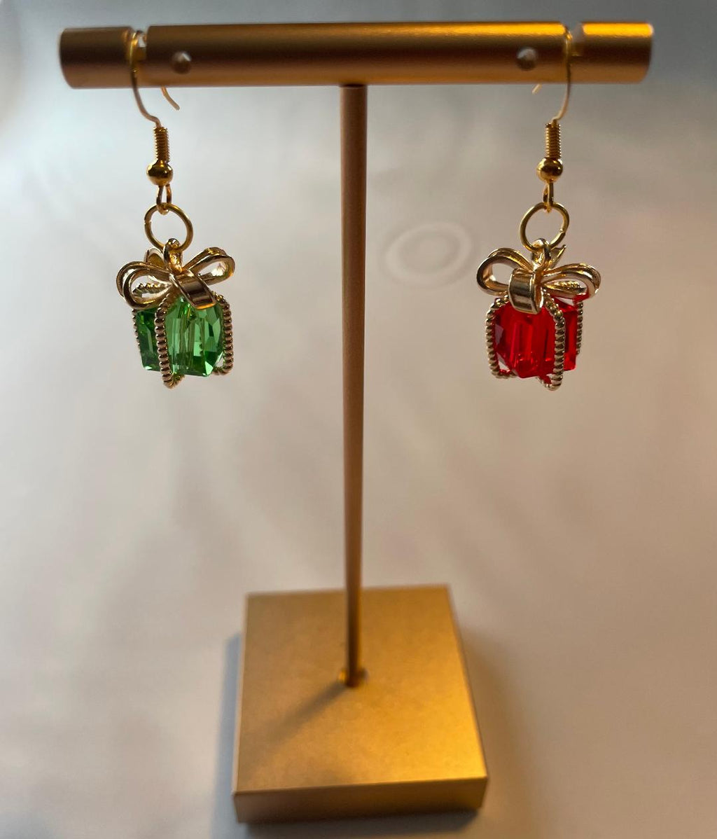 Gold bow-shaped earrings with red and green gemstones on a gold stand against a white background